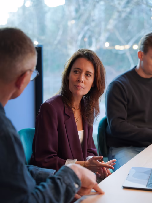 Twee mannen en een vrouw hebben een gesprek rond een tafel in een kantooromgeving met een laptop zichtbaar.