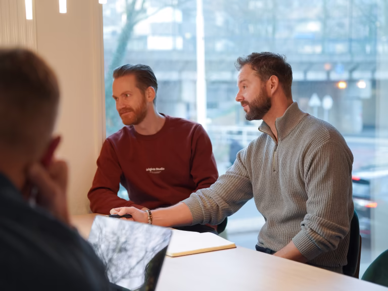 Twee mannen zittend aan een tafel in een gesprek tijdens een vergadering, met een raam op de achtergrond.