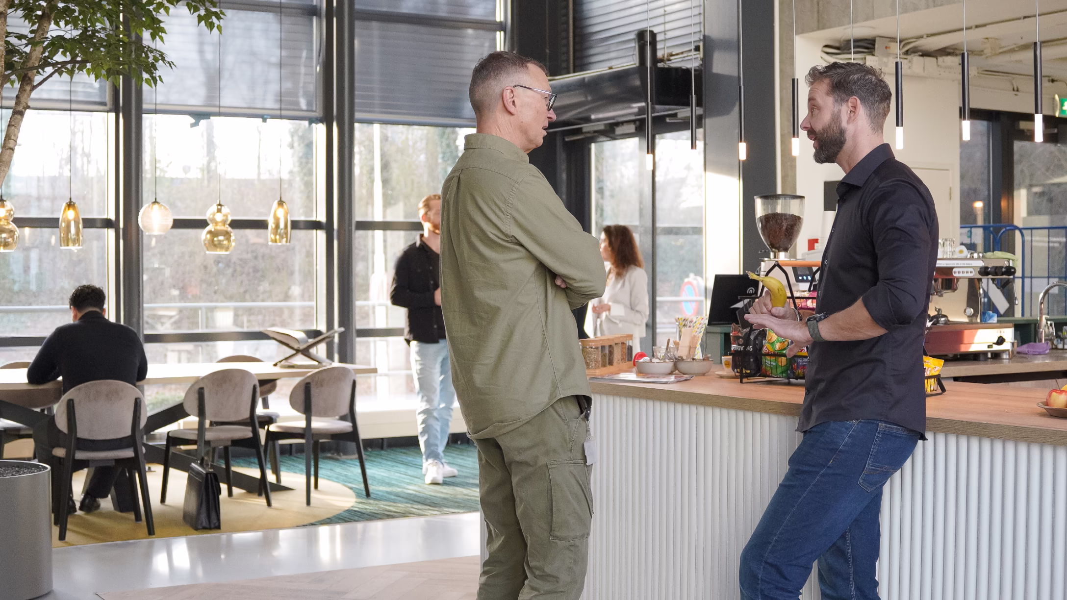 Twee mannen voeren een gesprek bij een bar in een moderne, lichte kantoorruimte met ramen van vloer tot plafond.