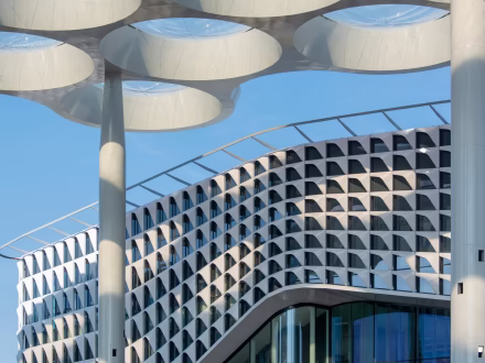 Modern gebogen gebouw met geometrische gevel en grote pilaren met ronde openingen tegen een blauwe lucht.