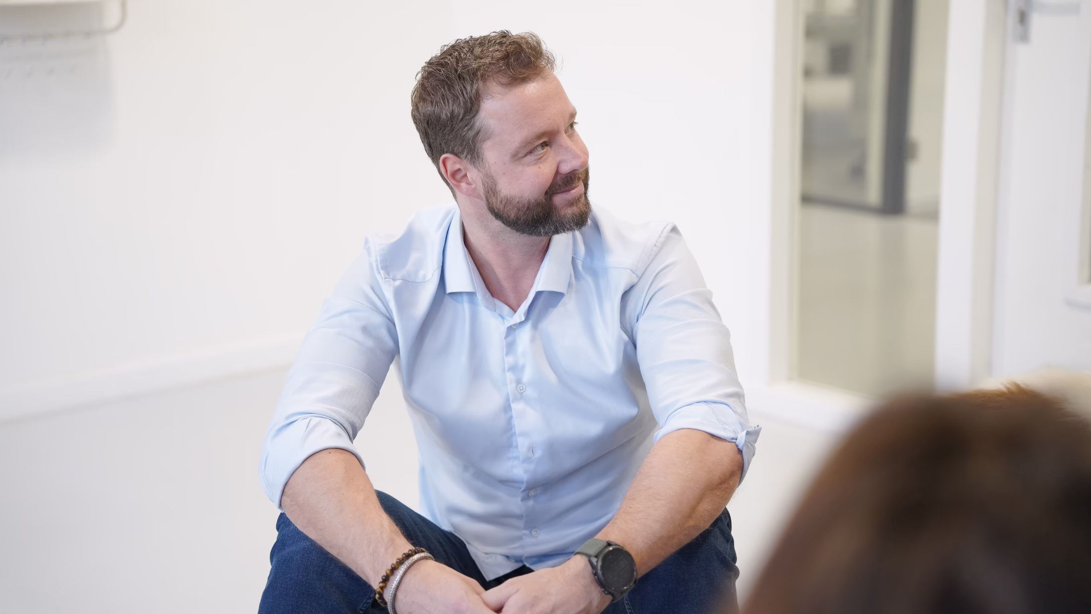 Man met baard en lichtblauw overhemd kijkt glimlachend naar rechts zittend in een lichte kamer.