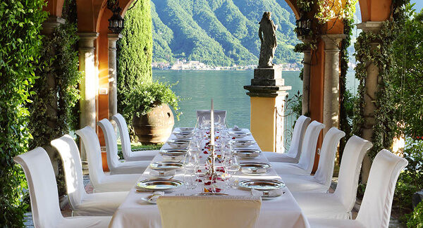 Long outdoor dining table set with white cloth and elegant tableware, surrounded by white chairs, overlooking a lake and green mountains.