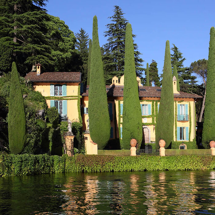 Yellow villa with green shutters surrounded by tall cypress trees on a lakeshore with a small covered boat docked in front.