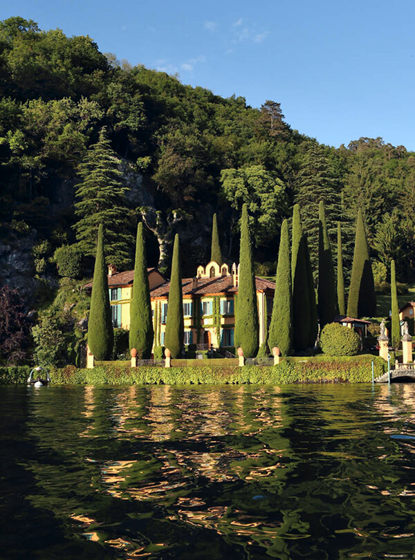 Modern outdoor pool with wooden deck, lounge chairs under white umbrellas, red foliage, and a mountain backdrop.