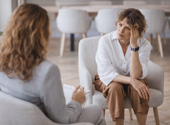 A distressed woman with curly hair sitting in a chair holding her head, talking to another person taking notes in an office setting.