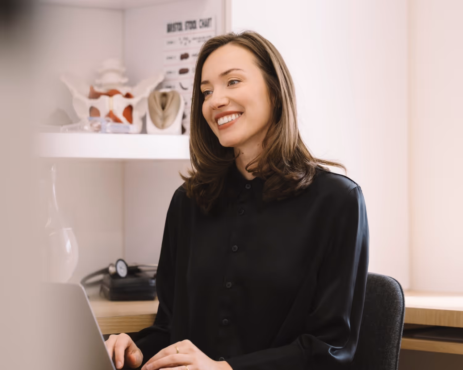 Smiling woman with brown hair sitting at a desk working on a laptop in a bright office.