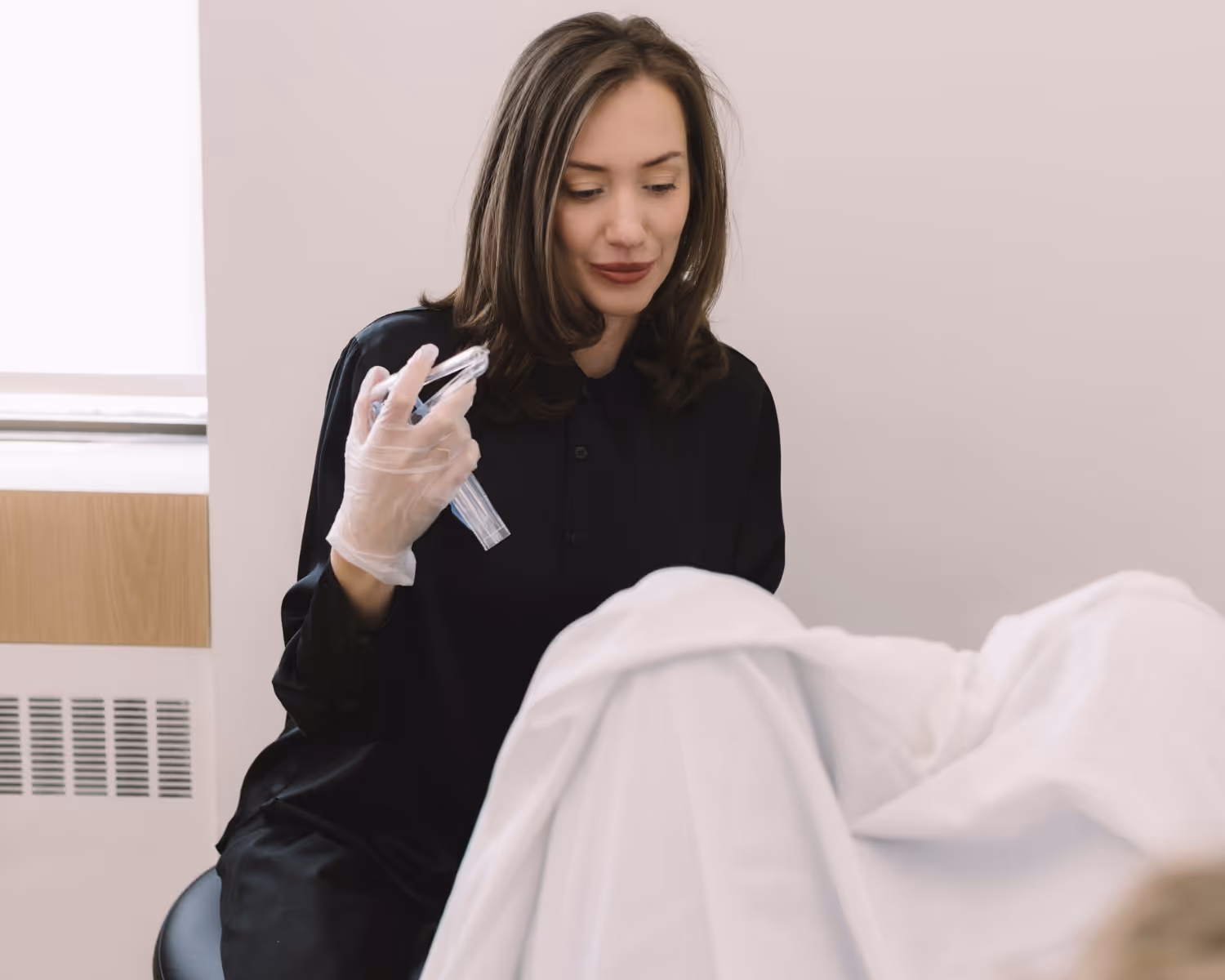 Medical professional wearing gloves holding a speculum, preparing for a gynecological exam with a patient covered by a white sheet.