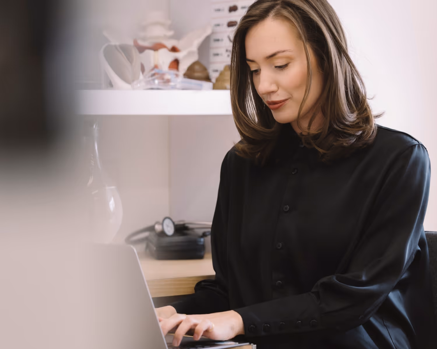 Woman in black blouse typing on a laptop with medical models and equipment in the background.