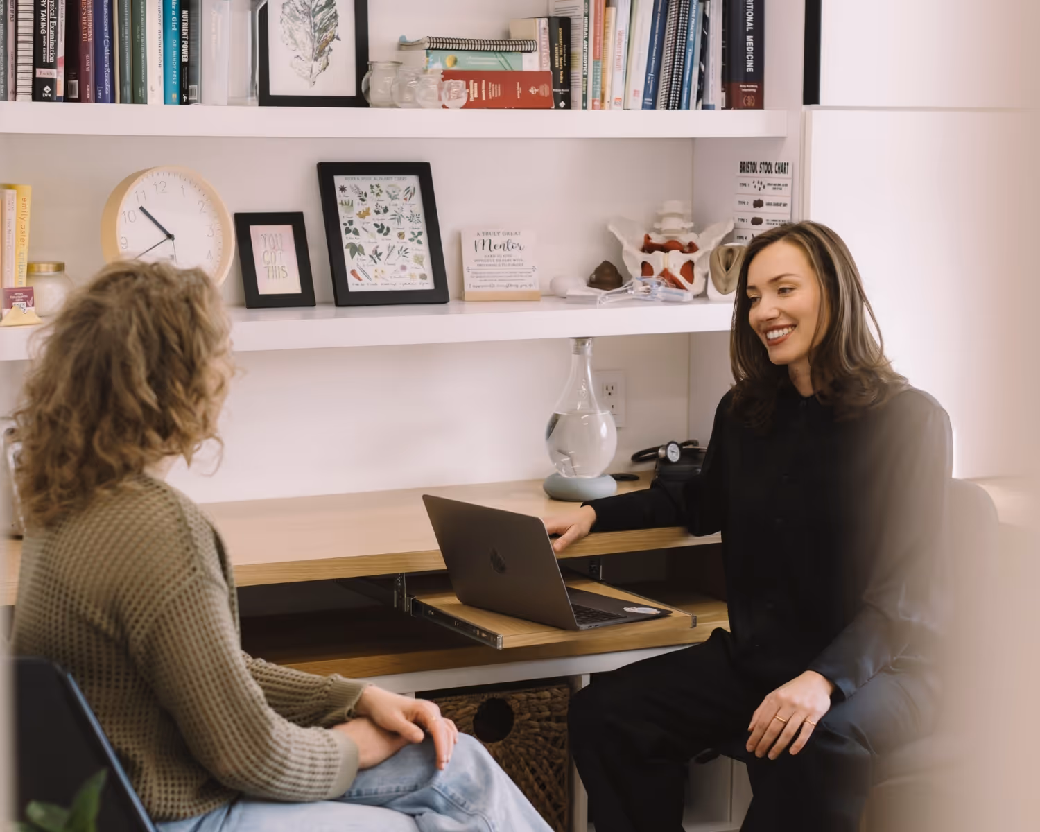Two women in an office with bookshelves, one seated with a laptop, smiling and talking.