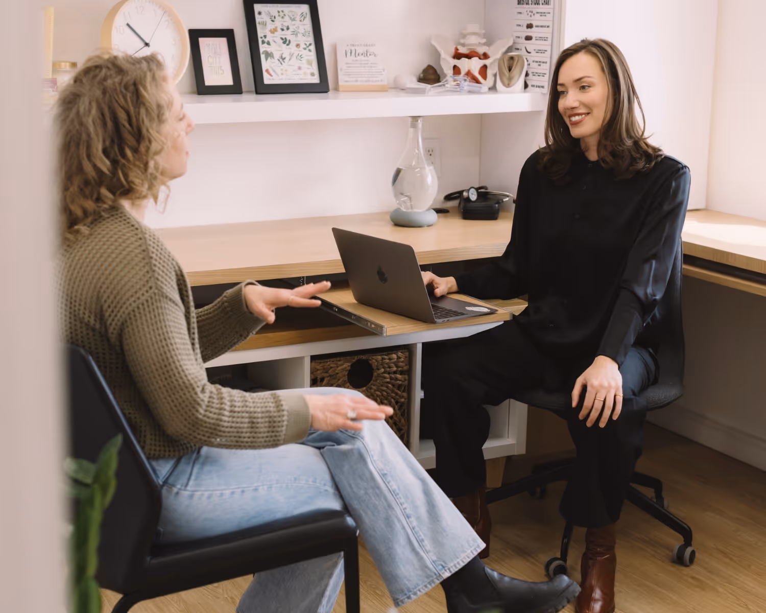 Two women having a friendly discussion in a bright office, one using a laptop on a wooden desk.