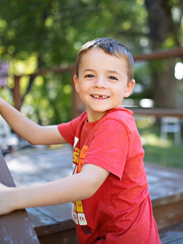 Smiling young boy with short brown hair wearing a red t-shirt outdoors.