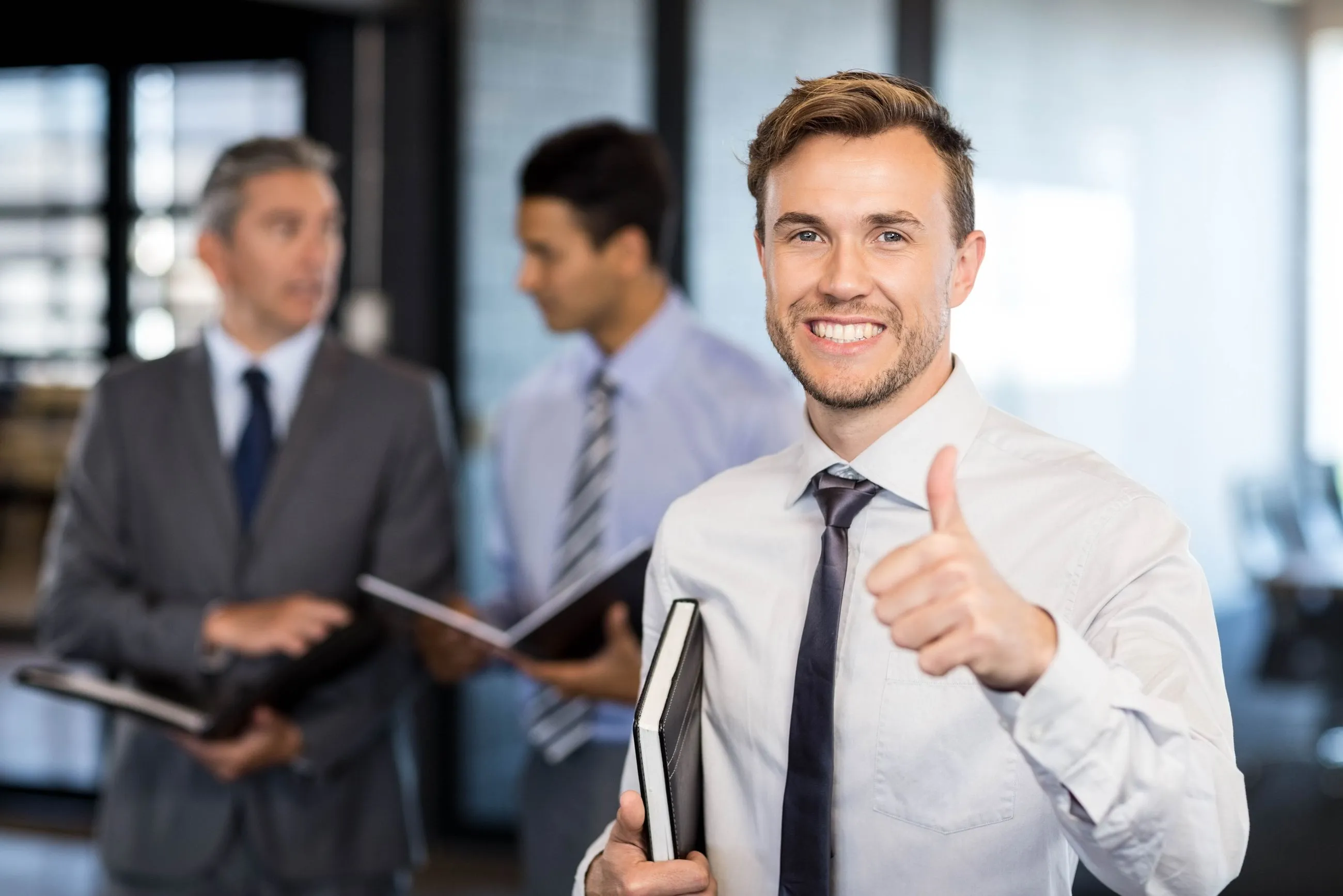 Smiling businessman in a shirt and tie giving a thumbs-up while holding a notebook, with two colleagues talking in the background.
