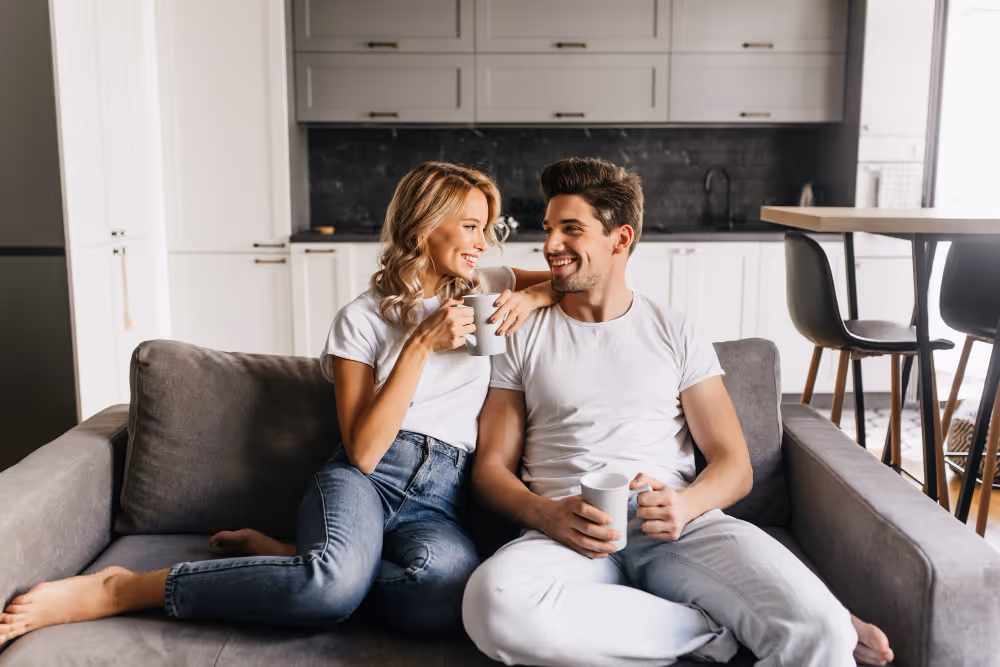 Smiling couple sitting close on a grey sofa in a modern kitchen, each holding a white mug.
