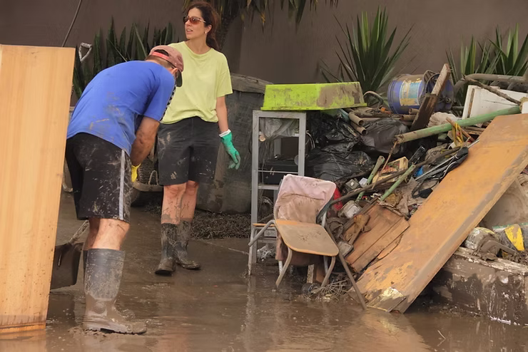 Two people wearing rubber boots and muddy clothes cleaning debris in a flooded area with piled-up damaged items in the background.