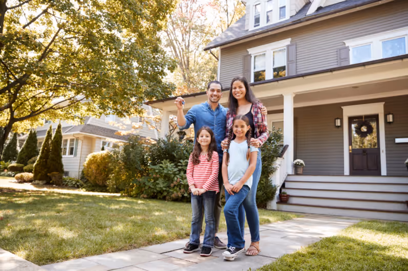 Smiling family of four standing on a walkway in front of a house with a man holding house keys.