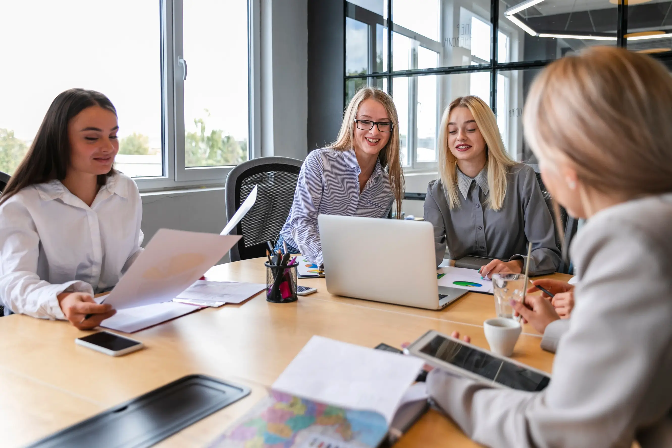 Four women in an office meeting around a table with papers, a laptop, and a tablet.