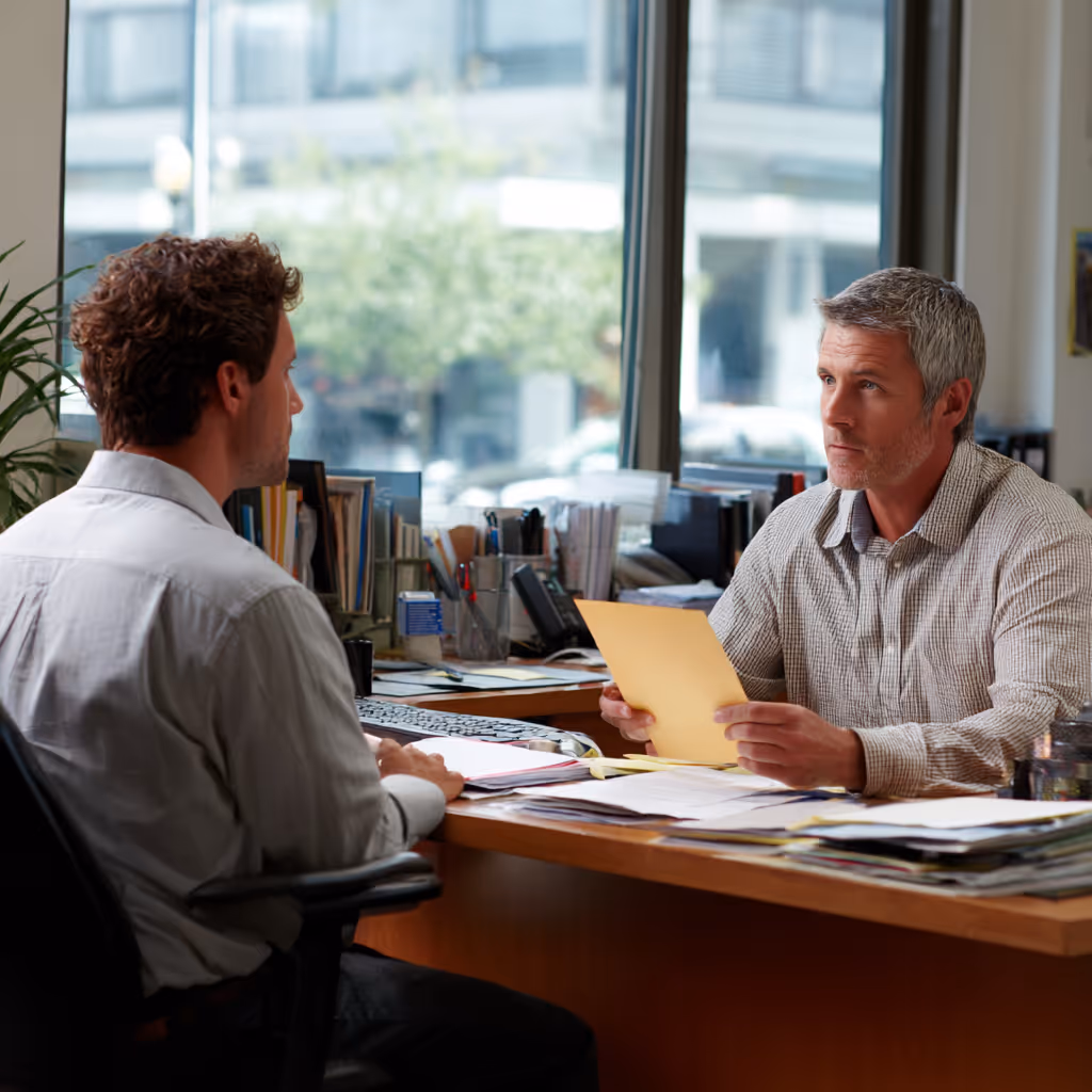 Two men having a serious discussion across an office desk with documents and files.