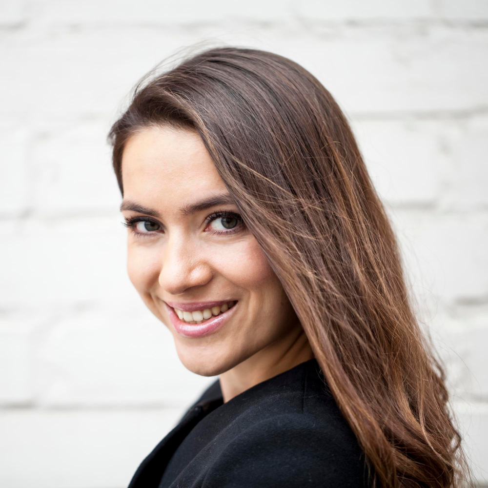 Young woman with long brown hair smiling over her shoulder against a white brick wall.