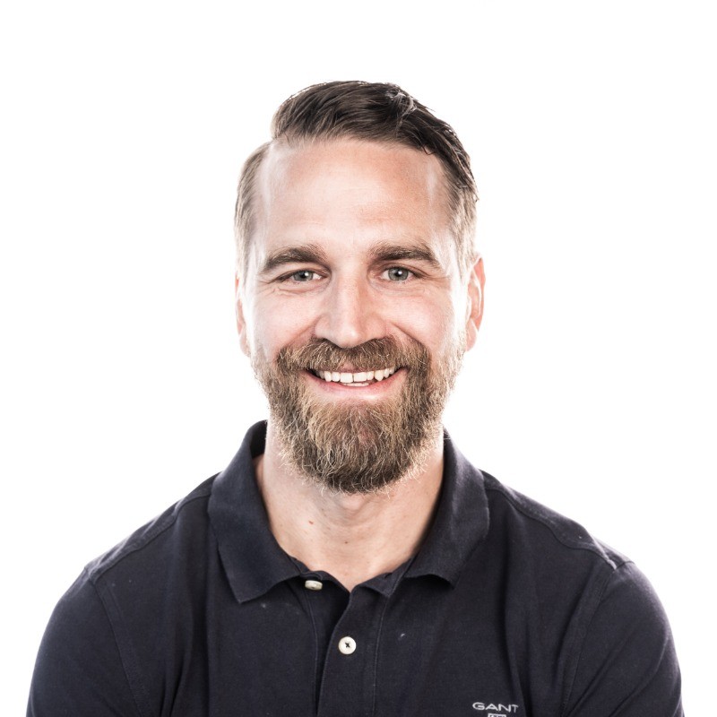Smiling man with brown hair and beard wearing a dark polo shirt against a white background.