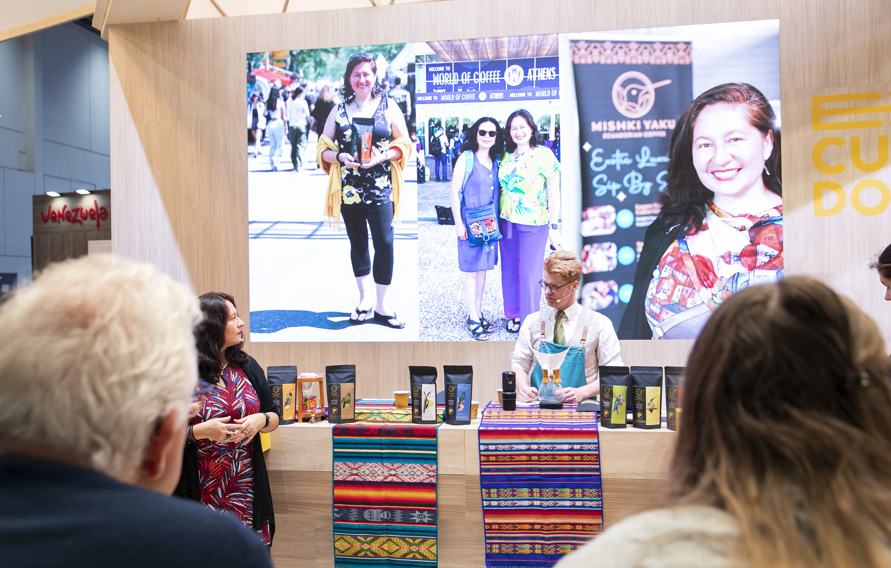 Man in apron prepares coffee at a colorful cloth-covered booth with coffee bags, a woman stands nearby, and a large screen behind them displays photos of smiling people and a coffee logo.