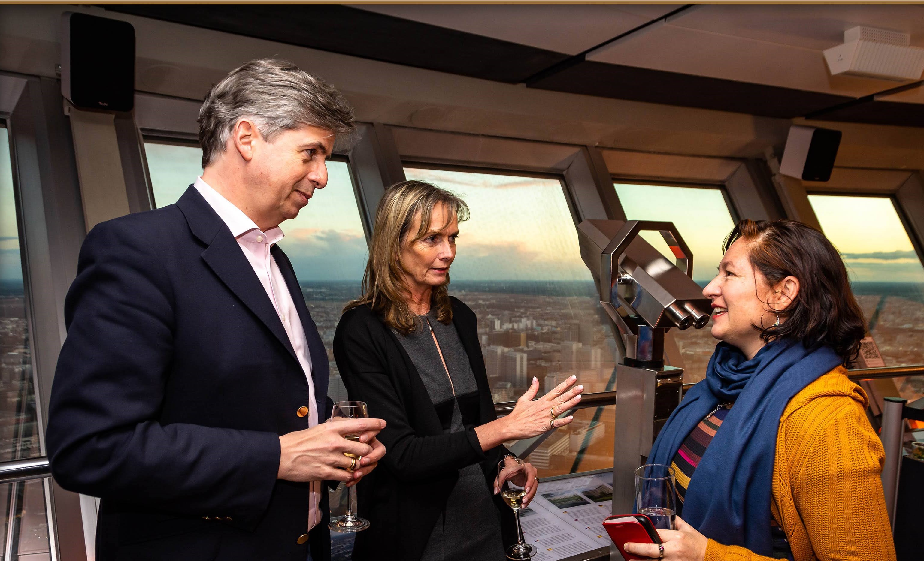 Three adults holding drinks and conversing inside a high-rise observation deck with city views through large windows.