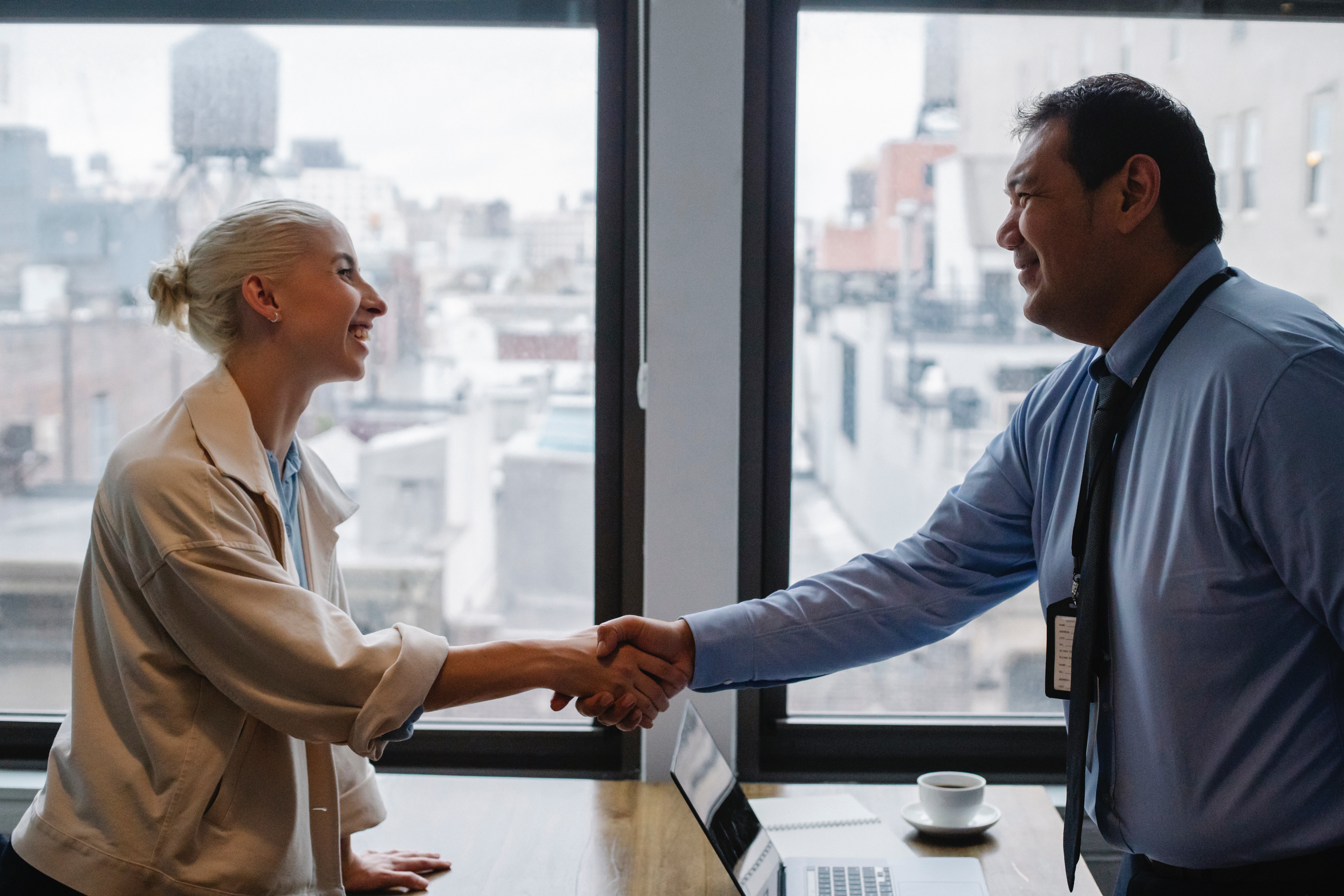 Two people smiling and shaking hands across a table with a laptop and coffee cup in an office with large windows overlooking city buildings.