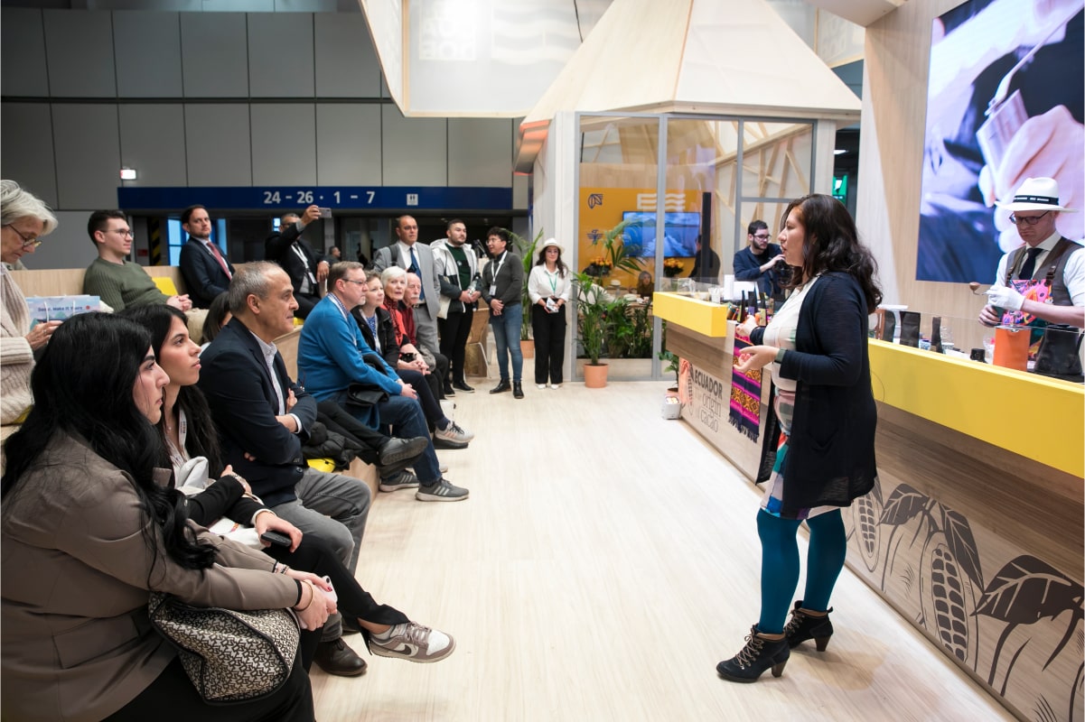 Woman giving a presentation to a seated and standing audience at an indoor event booth with Ecuadorian decorations.