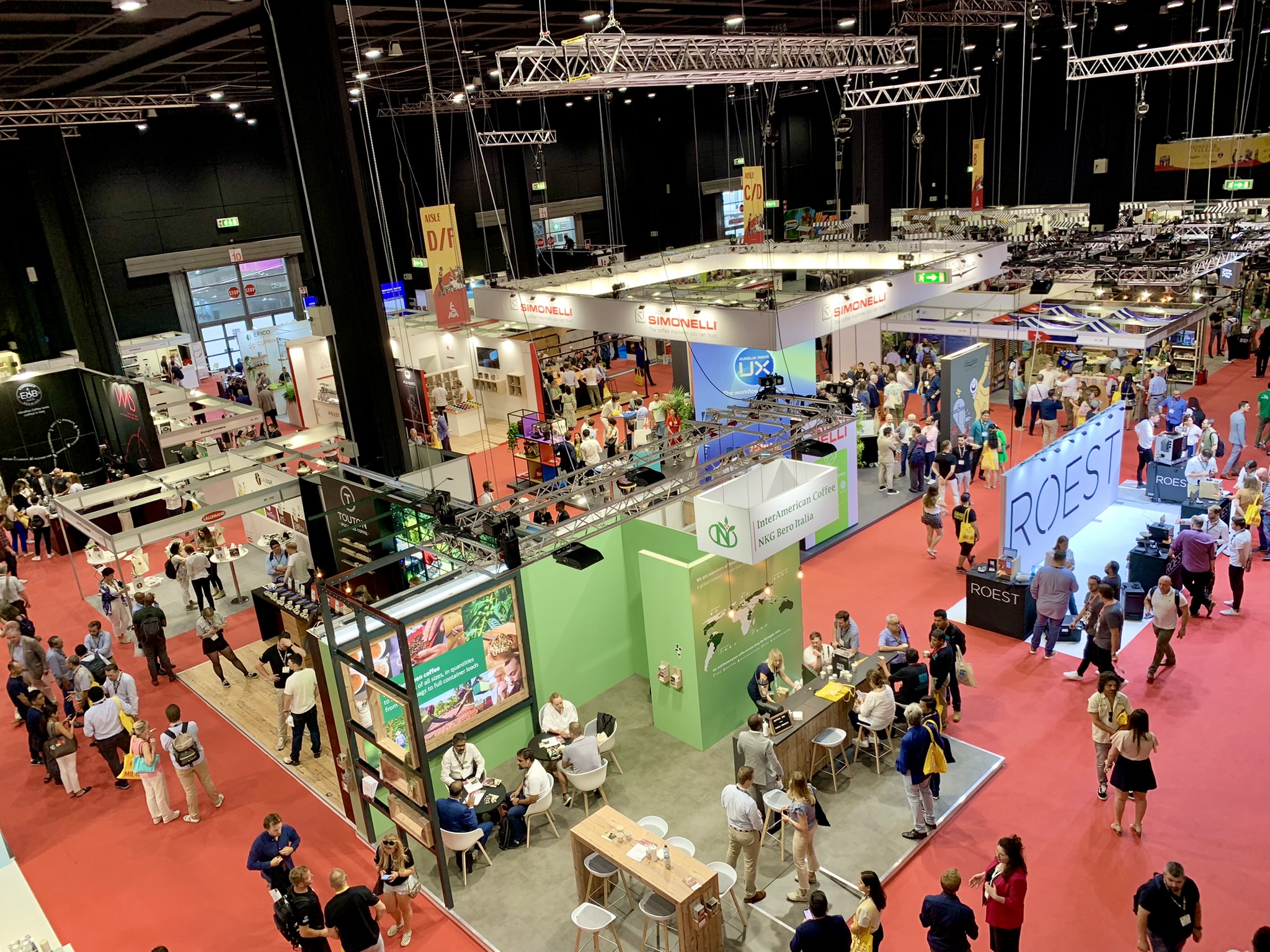 Overhead view of a busy trade show with multiple booths and crowds of people on red carpeted floor.