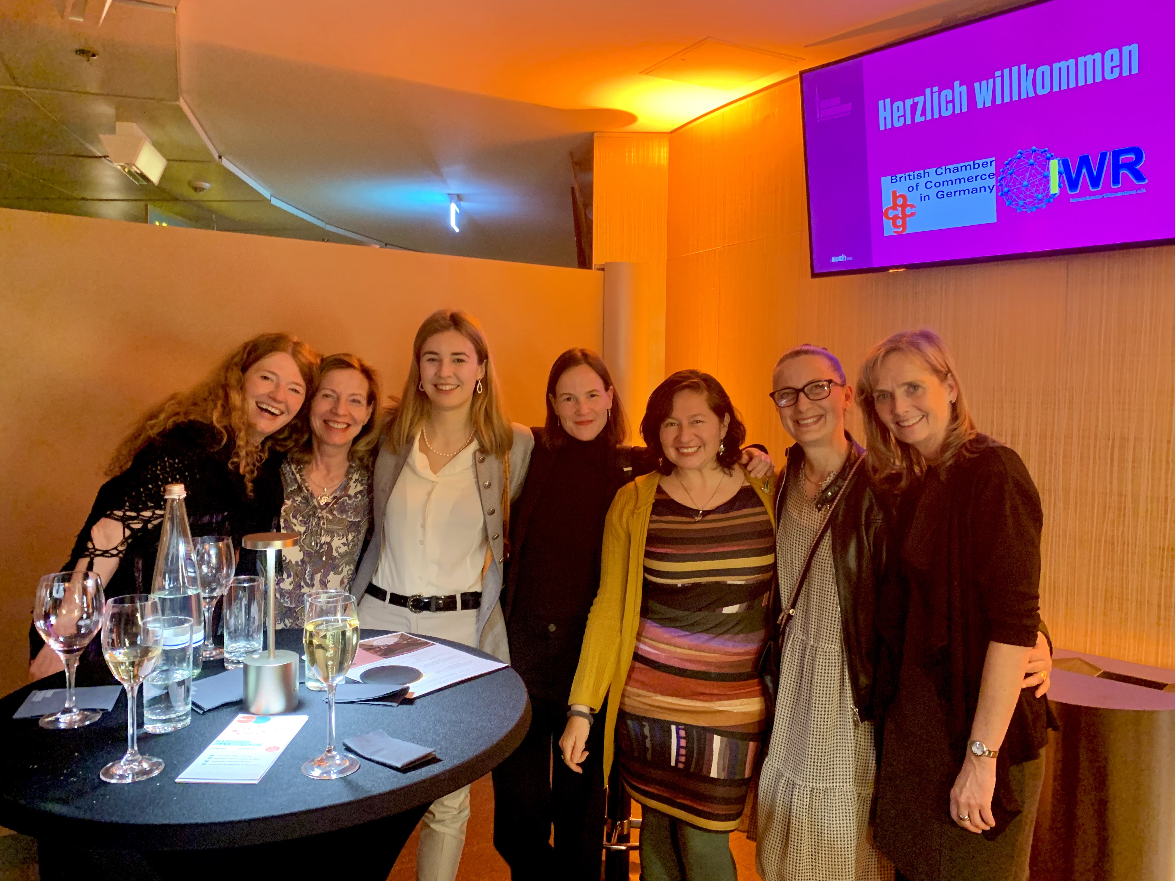 Seven smiling women standing closely together behind a table with glasses of water and wine, with a screen behind them showing a welcome message in German.