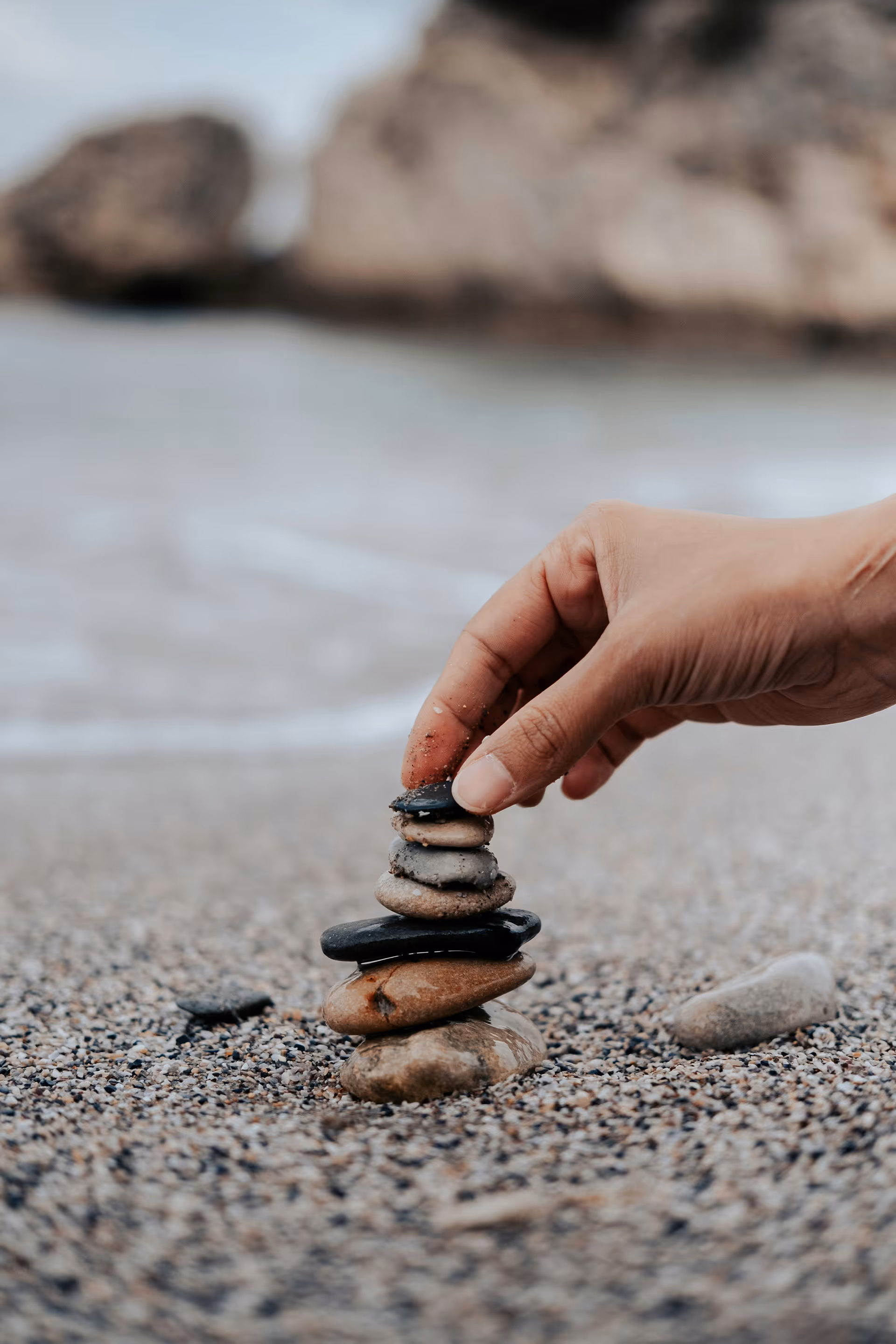 Hand stapelt kleine Steine am Strand aufeinander mit verschwommenem Felsen im Hintergrund.