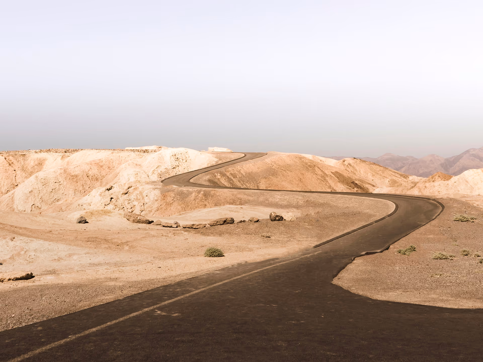 Kurvige Asphaltstraße führt durch trockene, bergige Wüstenlandschaft unter hellem Himmel.
