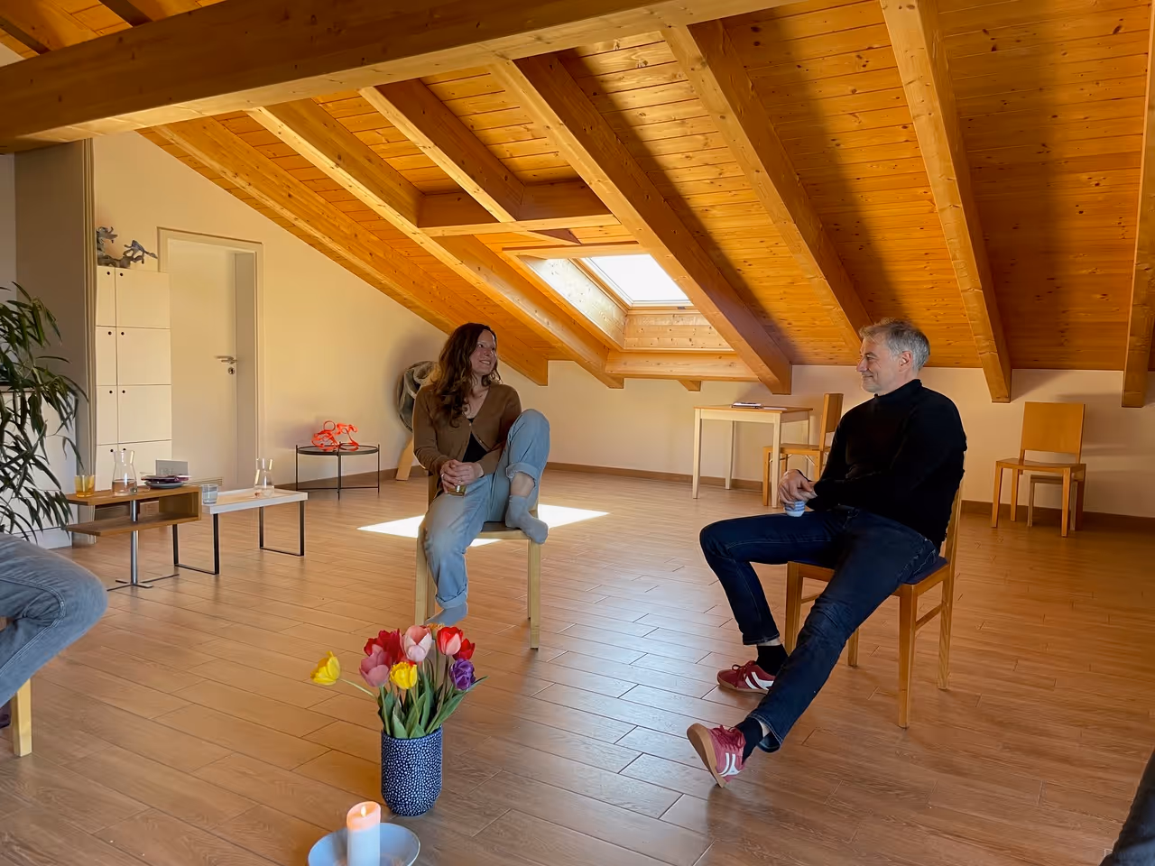 Zwei Personen sitzen auf Stühlen in einem Raum mit schräger Decke aus Holz, mit einem Fenster im Dach und einem Blumenstrauß auf dem Boden.