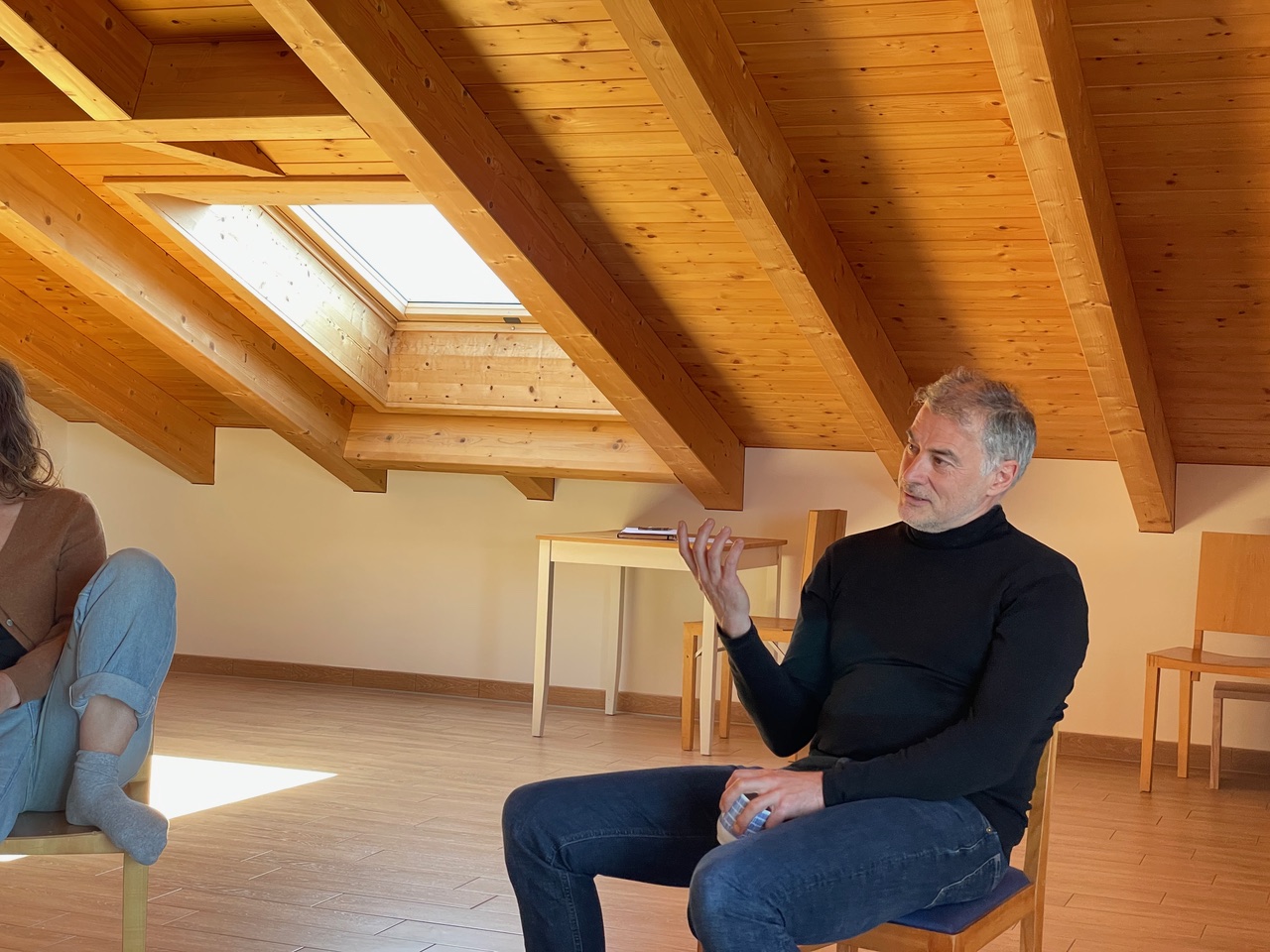 Mann im schwarzen Rollkragenpullover sitzt auf einem Stuhl in einem Raum mit schräger Holzdecke und spricht, während eine teilweise sichtbare Person rechts sitzt.