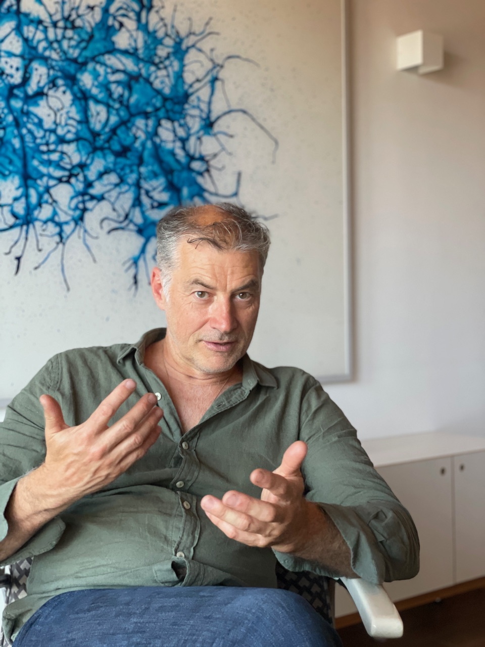 Mann in grünem Hemd sitzt und gestikuliert vor einem Bild mit blauem, abstraktem Muster an der Wand.