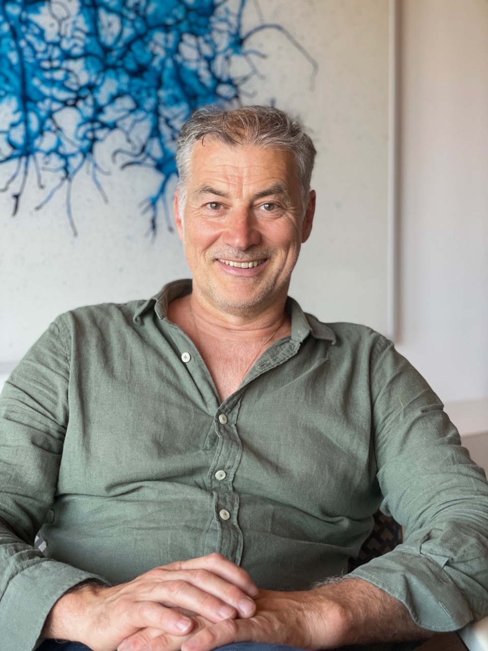 Lächelnder Mann mittleren Alters mit grauen Haaren trägt ein grünes Hemd und sitzt mit verschränkten Händen.