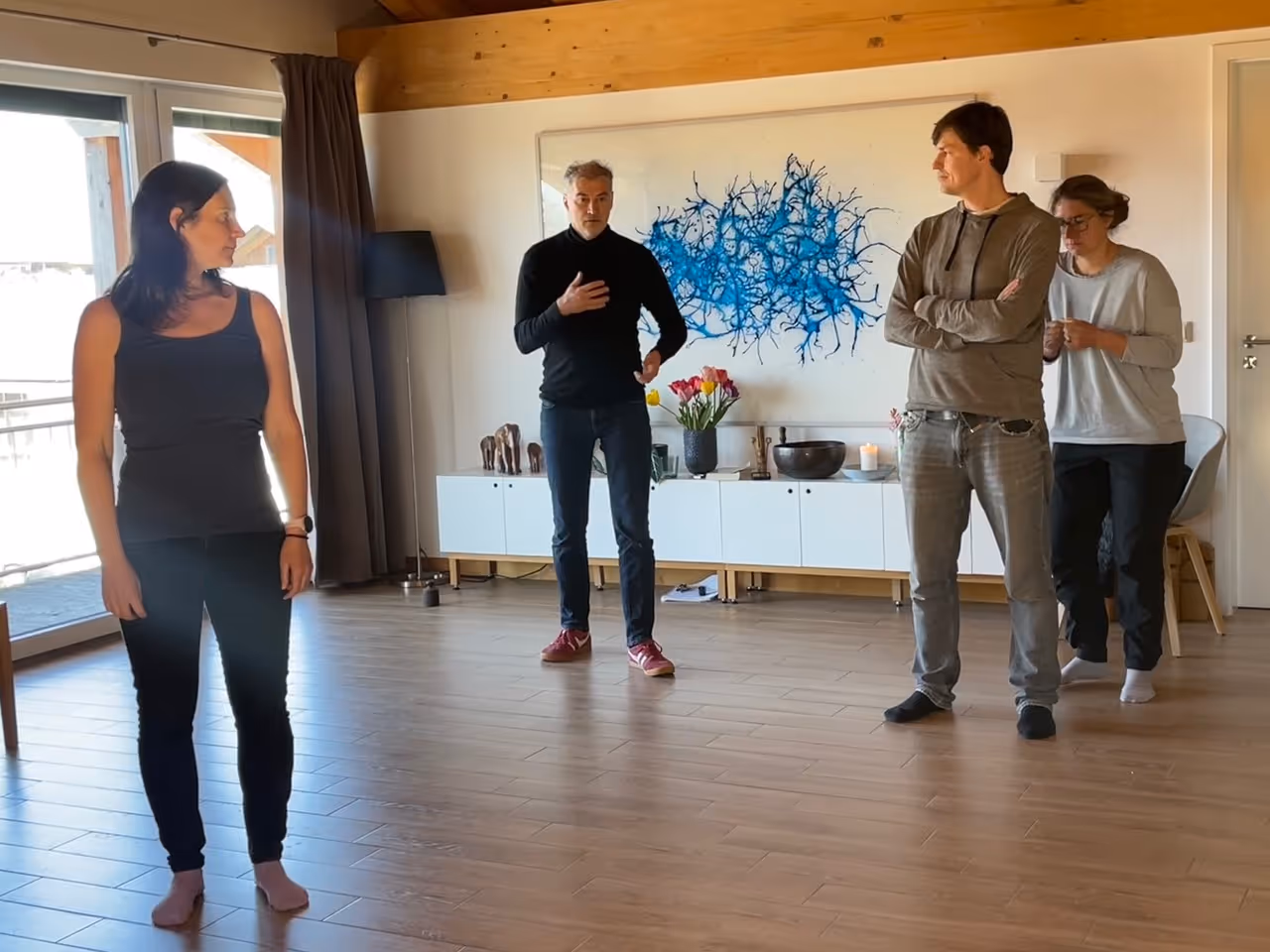 Vier Personen stehen in einem hellen Raum mit Holzboden, eine Frau schaut zur Seite, ein Mann spricht, während zwei weitere Personen an der Wand lehnen.