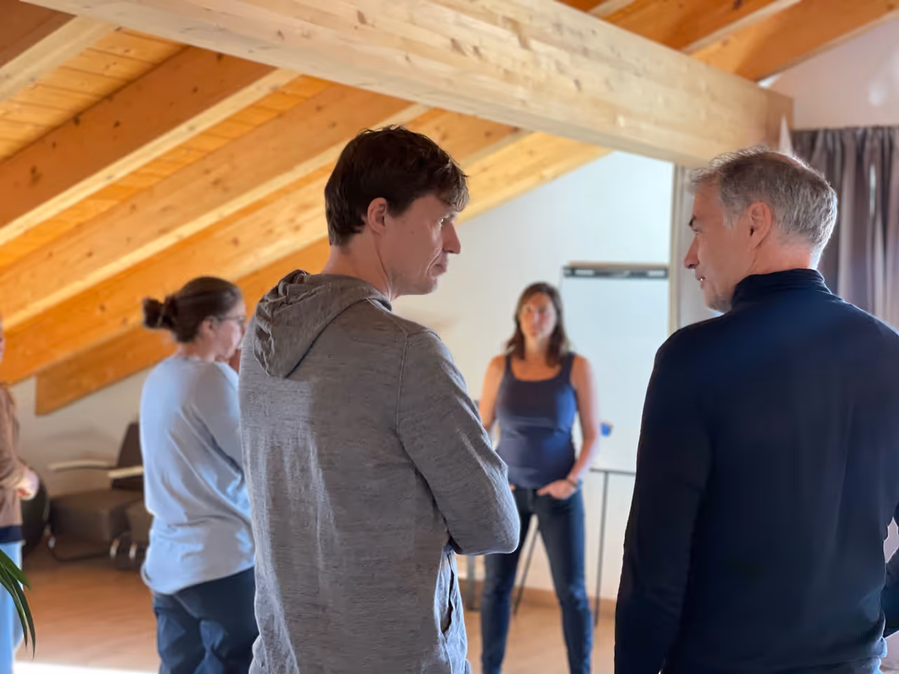 Vier Erwachsene in einem Raum mit sichtbaren Holzbalken, zwei Männer im Vordergrund im Gespräch, zwei Frauen im Hintergrund.