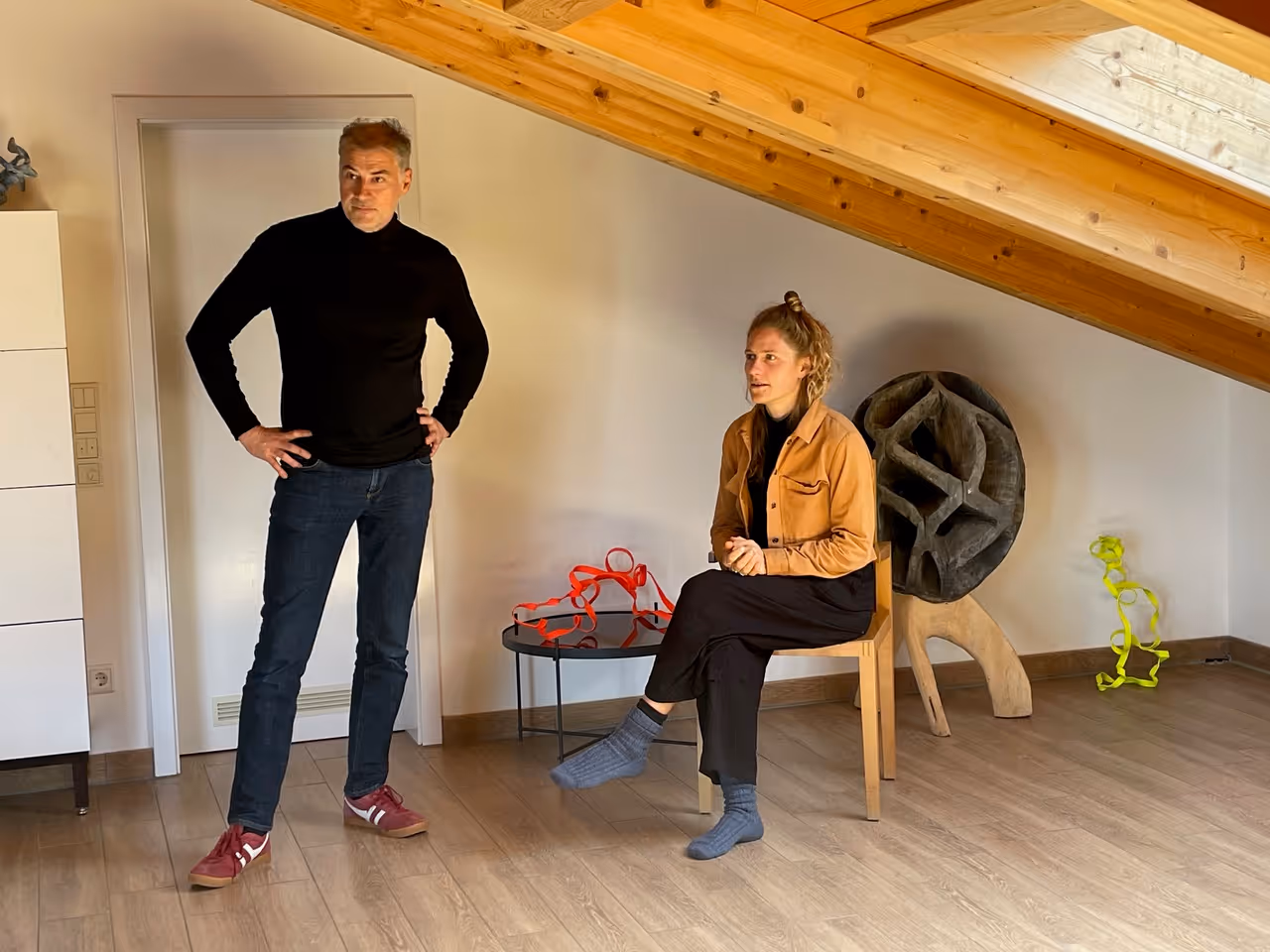 Ein Mann in schwarzem Rollkragenpullover und Jeans steht mit den Händen in der Hüfte, während eine sitzende Frau in brauner Jacke und schwarzen Hosen in einem Raum mit Holzdecke und moderner Kunst sitzt.