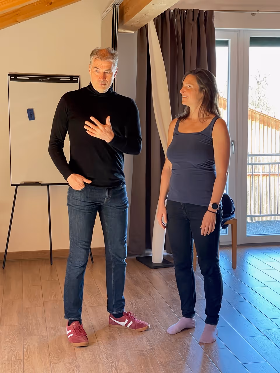 Ein Mann in schwarzem Rollkragenpullover und Jeans spricht, während eine Frau in ärmellosem Top und Jeans barfuß daneben steht und lächelt.