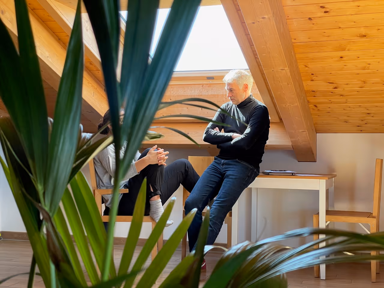 Zwei Menschen im Gespräch in einem Dachzimmer mit Holzbalken, teilweise durch Zimmerpflanzen verdeckt.