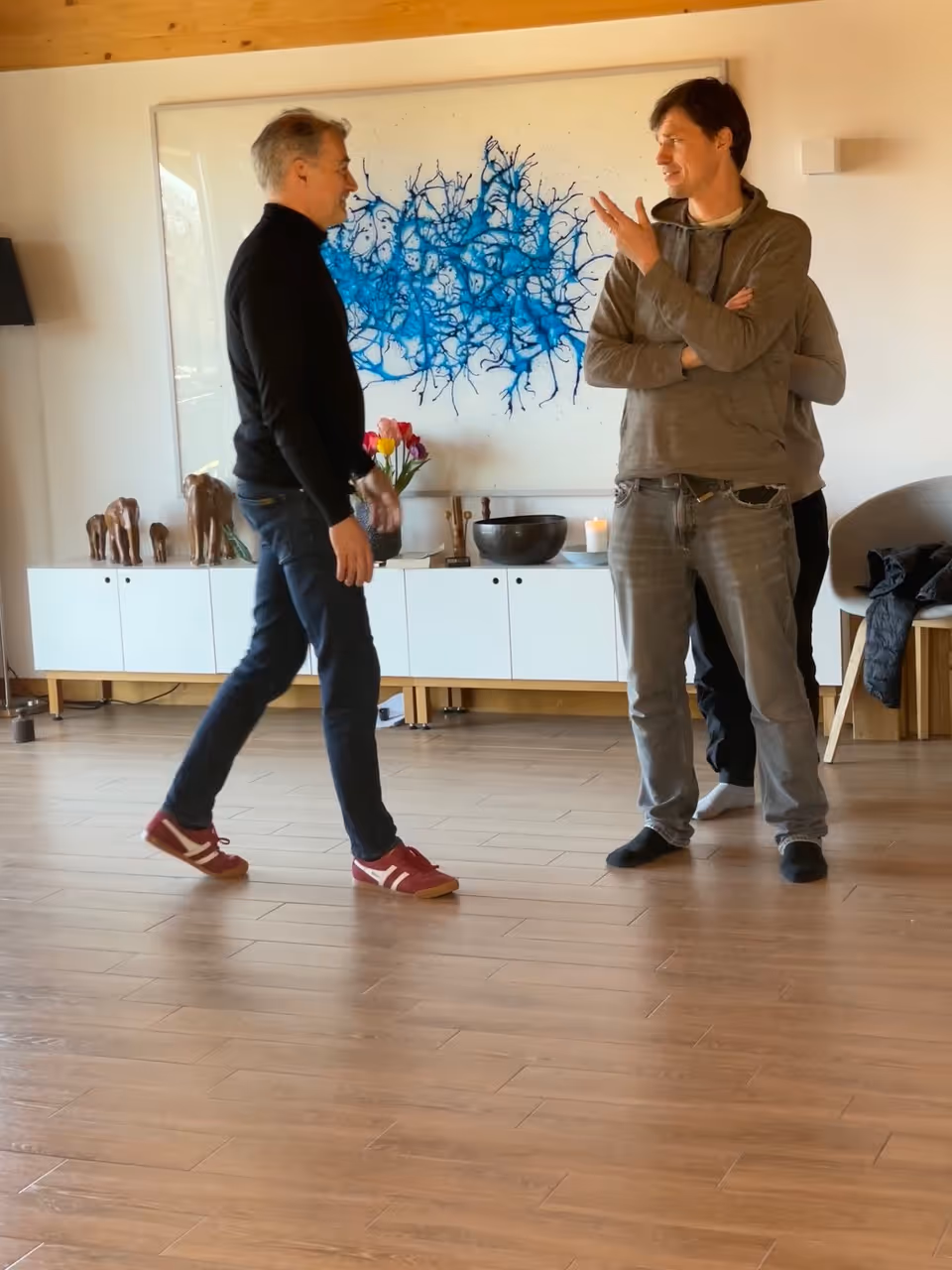 Zwei Männer im Gespräch in einem modernen Raum mit Holzboden und einem blauen abstrakten Gemälde an der Wand.