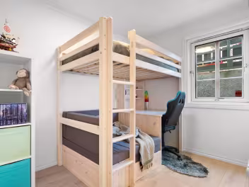 Barnerom med en trebunkeseng, skrivebord under øverste køye, kontorstol og vindu med utsikt til bygninger.