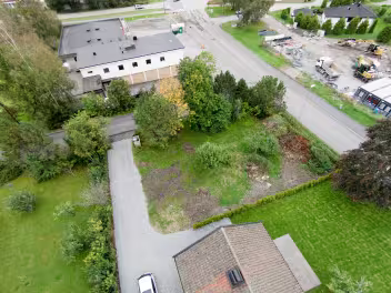 Luftfoto av en boligeiendom med en oppkjørsel, gressplen og trær, med en vei og flere bygninger i bakgrunnen.