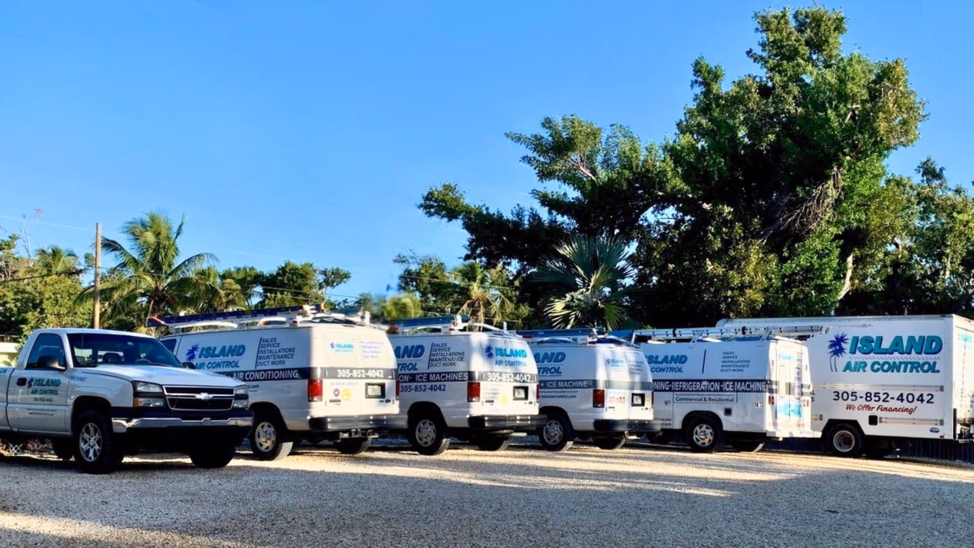 Fleet of white Island Air Control service trucks and vans parked on gravel with trees and clear blue sky in the background.