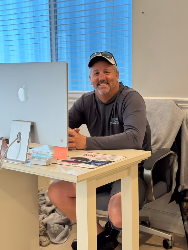 Island Air Control owner wearing a cap and sunglasses on his head, smiling while sitting at a desk with an iMac computer in an office.