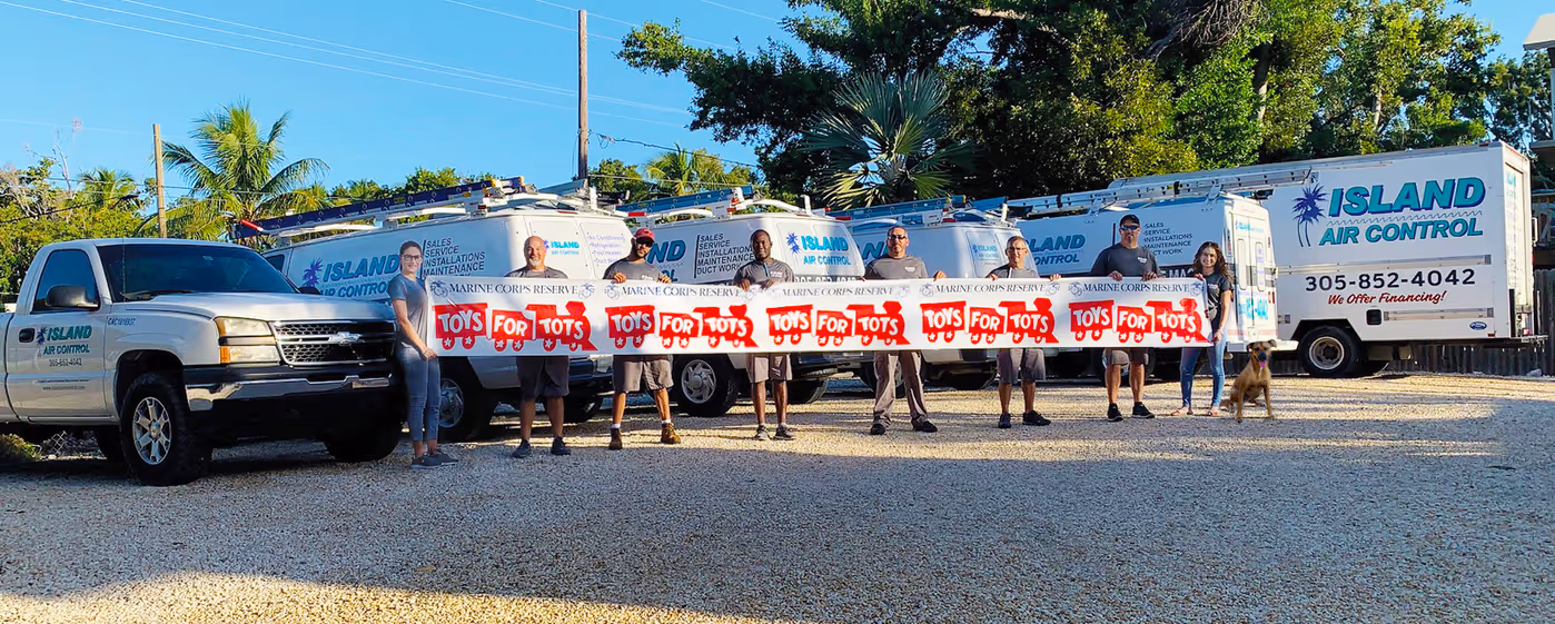 Group of people standing in front of Island Air Control service trucks holding a long Toys for Tots banner, with a dog sitting on the right side.