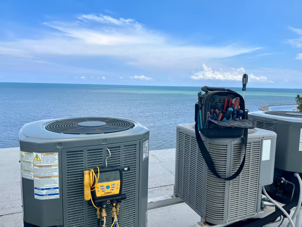 Three rooftop air conditioning units overlooking a calm ocean under a blue sky with scattered clouds, one unit has a tool bag with hand tools on top.