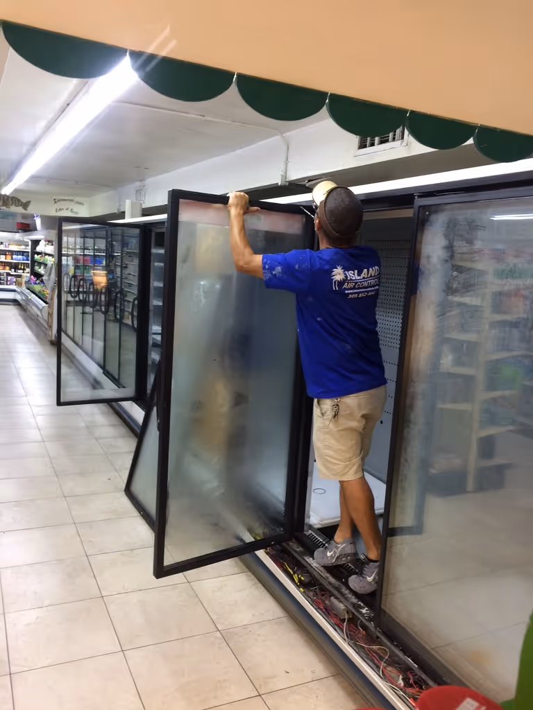 Technician in blue Island Air Control shirt repairs or installs a glass door on a large commercial refrigerator in a store aisle.