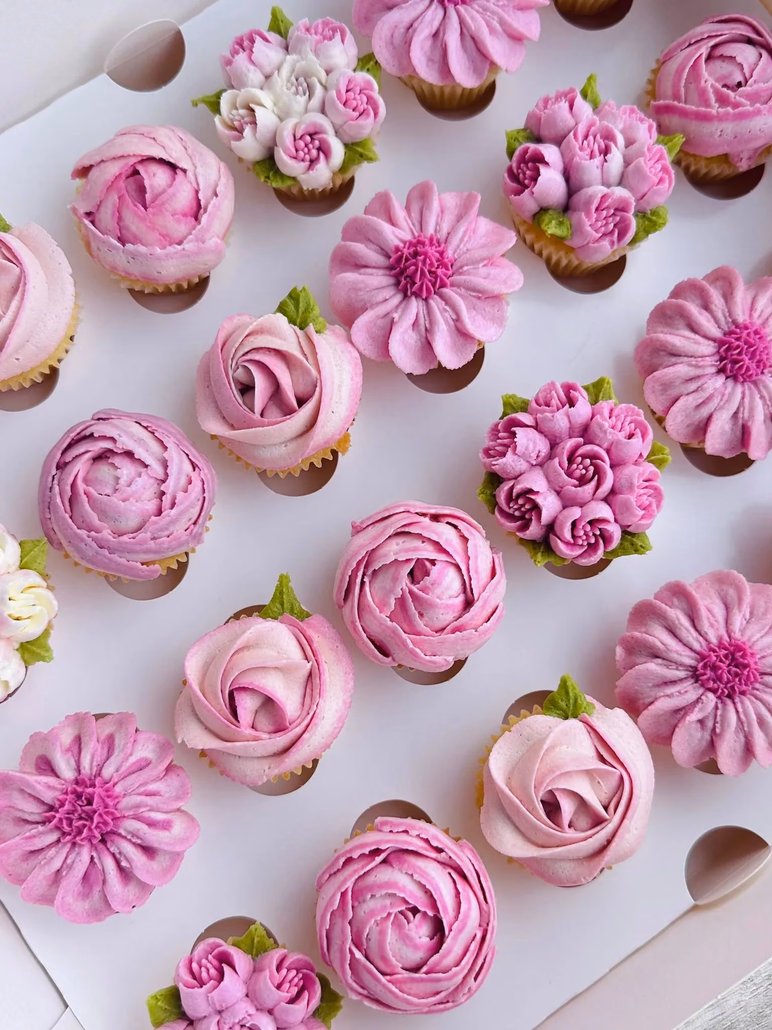 Box of assorted cupcakes decorated with pink and white floral buttercream frosting resembling roses and other flowers.