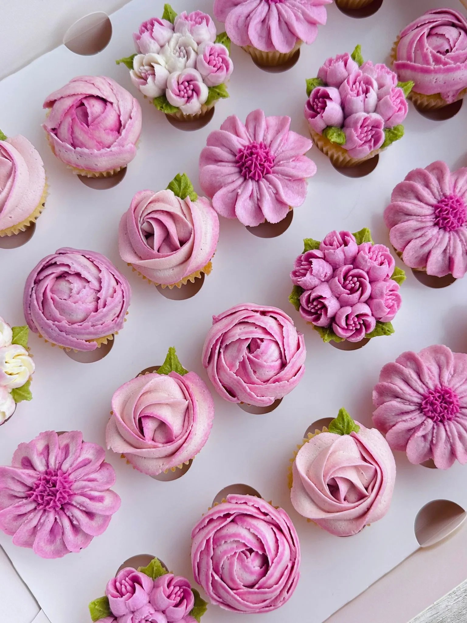 Box of assorted cupcakes decorated with pink and white floral buttercream frosting resembling roses and other flowers.